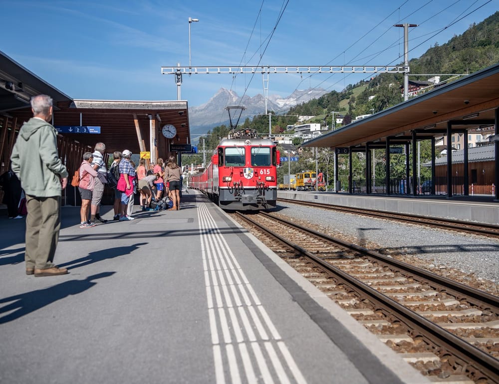 Rhb fährt in Bahnhof rein, Passagiere warten auf der Linken Seite auf dem Perron