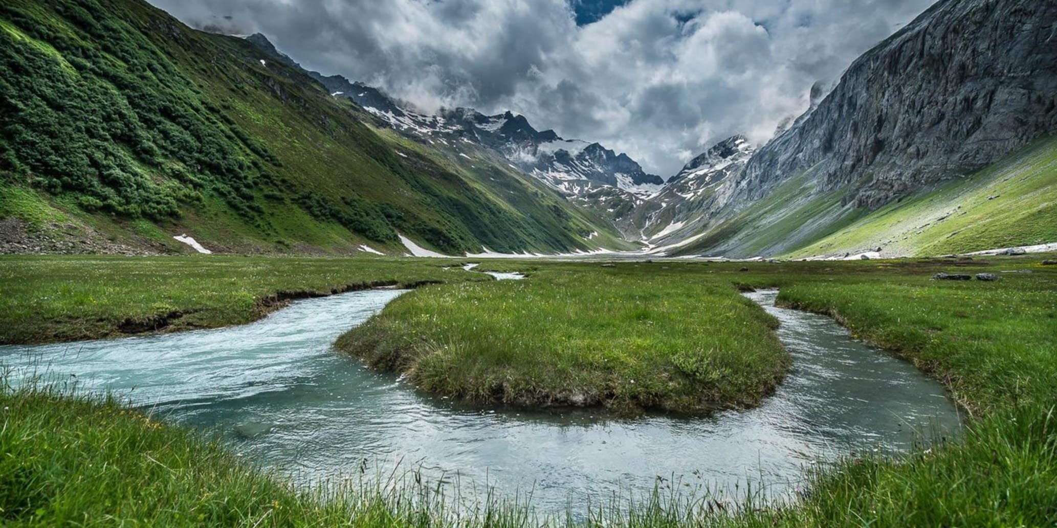 Bach auf Hochebene inmitten von grüner Wiese umrahmt von Bergen