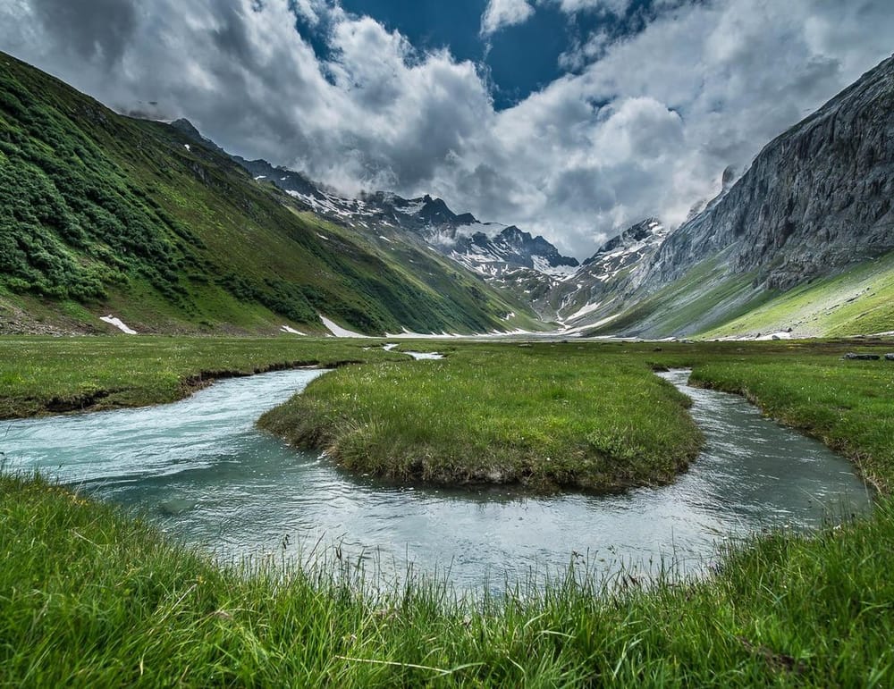 Bach auf Hochebene inmitten von grüner Wiese umrahmt von Bergen