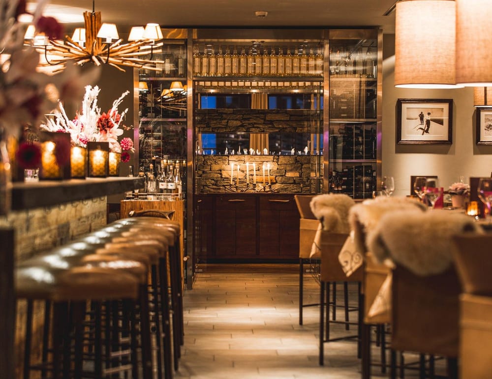 Bar warm beleuchtet, links Barhocker und Theke und rechts Hochstühle und Apérotische