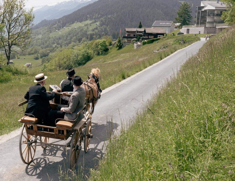 Pferdewagen auf kleiner Strasse mit mehreren Personen fahren vom Betrachter weg, im Hintergrund Dorf im Hochsommer