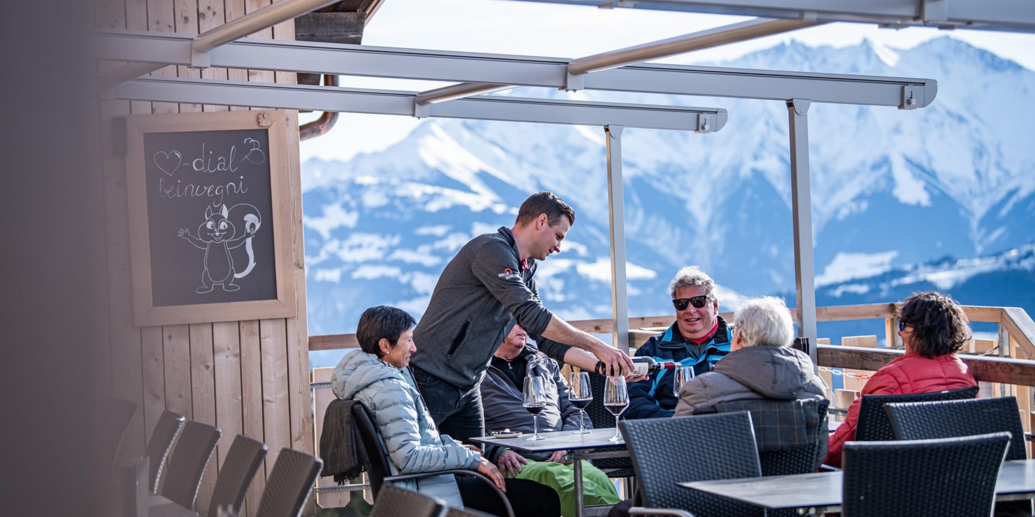 Terrasse mit Kellner und Gästen, es wird Rotwein serviert, im Hintergrund sind Berge