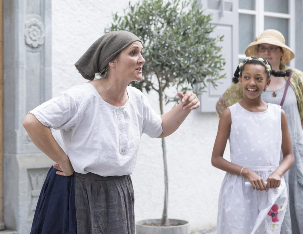 Szene aus Theaterstück, Frau mit Kopftuch und historischer Bauernkleidung daneben zwei weitere Personen welche sie anschauen