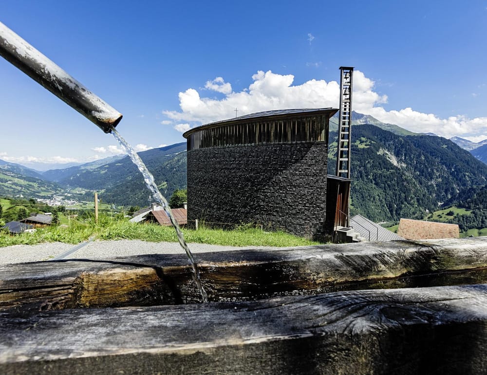Brunnen, im Hintergrund moderne halbrunde Holzkirche mit Bergen im Sommer
