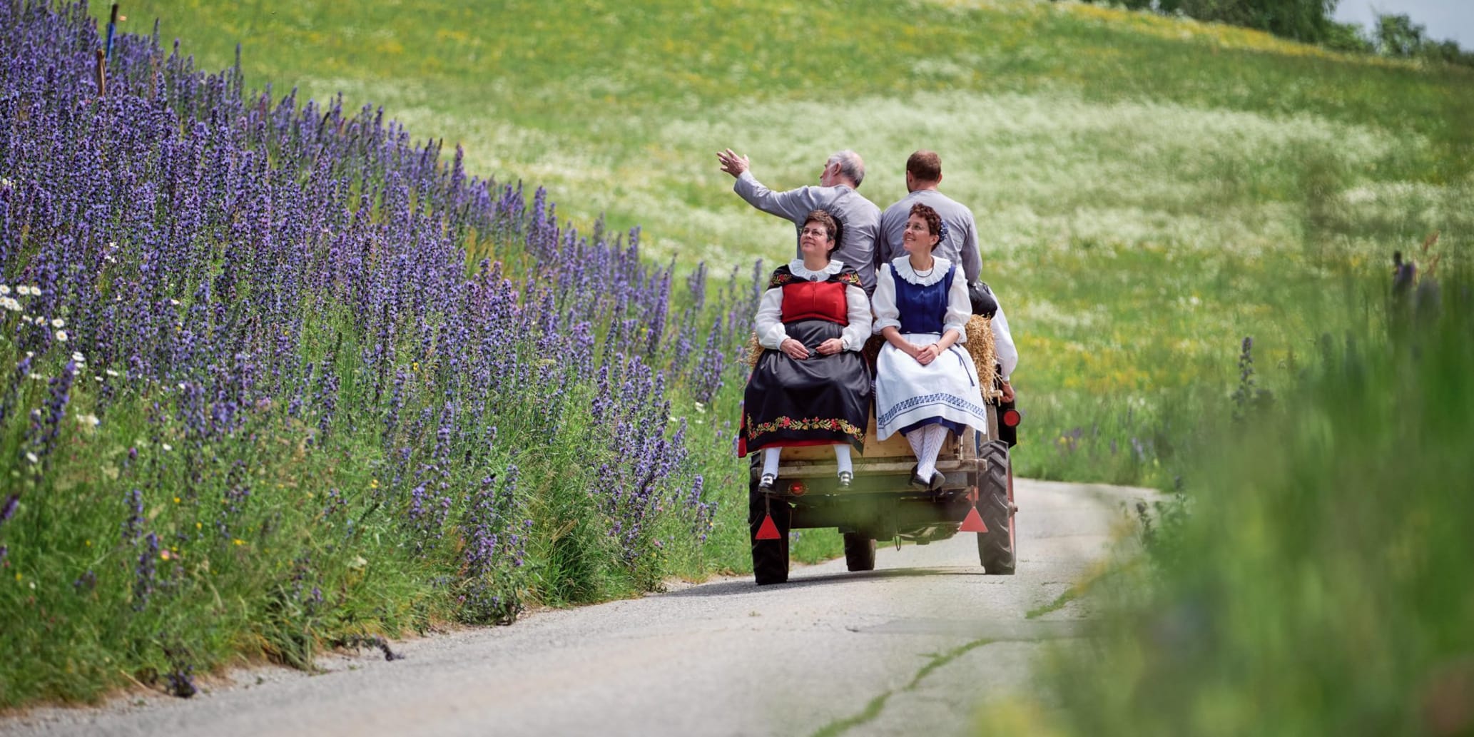 Rapid fährt mit zwei Landfrauen hintendrauf auf kleiner Strasse vom Betrachter weg. Links und rechts sind Blumenfelder.