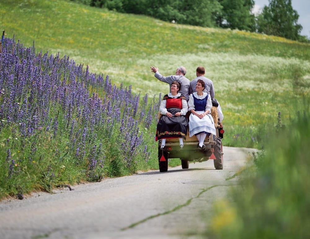 Rapid fährt mit zwei Landfrauen hintendrauf auf kleiner Strasse vom Betrachter weg. Links und rechts sind Blumenfelder.